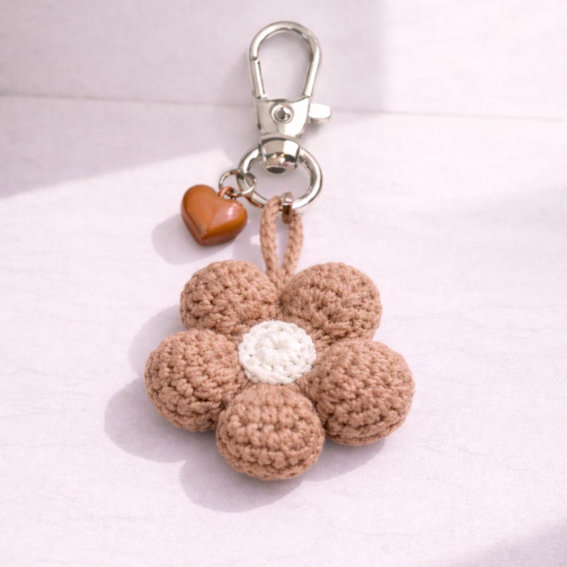 May include: A handmade keychain with a crocheted flower design. The flower has five brown petals and a white centre. A small brown heart charm hangs from the silver clasp. Ideal for keys or bags.