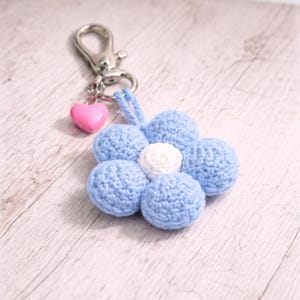 May include: A blue crochet flower keyring with a white central detail and a pink heart charm. The keyring has a silver clasp. The flower is made of soft yarn. The item is suitable for attaching to keys or bags.