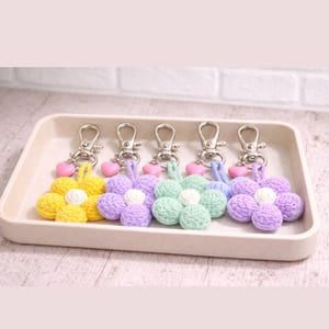 May include: Five crochet flower keyrings in yellow, purple, and green, each with a white centre and a silver clasp. Pink heart charms dangle from the clasps. The keyrings are displayed in a rectangular tray.