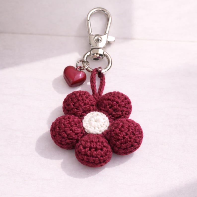 May include: A burgundy crochet flower keyring with a white centre and a small red heart charm. The keyring has a silver clasp for easy attachment to keys or bags. The flower design adds a touch of handmade charm.