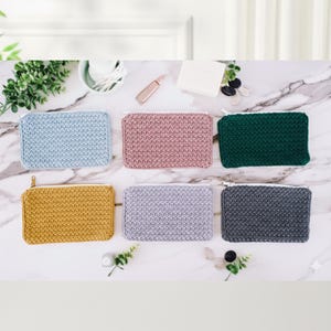 K&ouml;nnte beinhalten: Sechs rechteckige H&auml;keltaschen mit Rei&szlig;verschluss in verschiedenen Farben: Hellblau, Rosa, Gr&uuml;n, Gelb, Lavendel und Anthrazitgrau. Die Taschen sind auf einer Marmoroberfl&auml;che angeordnet und zeigen ihr strukturiertes Stichdesign und wei&szlig;e Rei&szlig;verschl&uuml;sse.