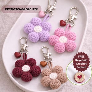 May include: Four handmade crochet flower keychains in shades of purple, pink, burgundy, and tan, each with a silver clasp and a heart charm. The keychains are arranged on a white tray. Text on the image reads "INSTANT DOWNLOAD | PDF" and "FREE Keychain Crochet Pattern".