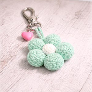 May include: A crochet flower keyring with a silver clasp and a pink heart charm. The flower has mint green petals around a white centre. A delightful handmade accessory.
