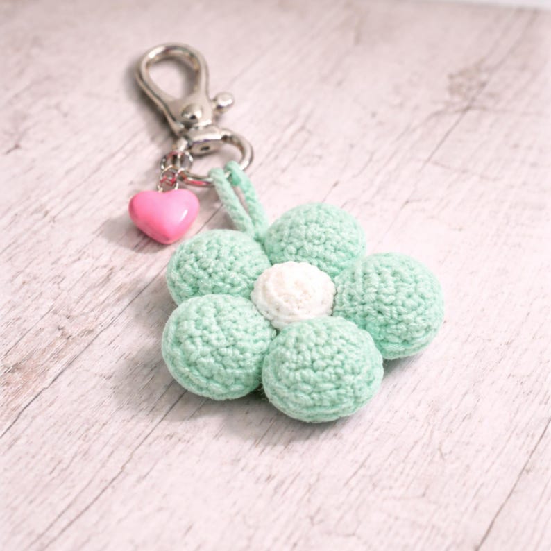 May include: A crochet flower keyring with a silver clasp and a pink heart charm. The flower has mint green petals around a white centre. A delightful handmade accessory.