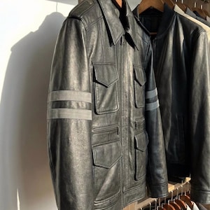 May include: Black leather jacket with four front pockets, a collared neckline, and gray stripes on the sleeves. The jacket is hanging on a wooden hanger. Another black jacket is visible in the background.