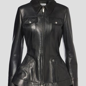 Könnte beinhalten: Schwarze Lederjacke mit Reißverschluss, zwei Brusttaschen und einem strukturierten Schößchen. Die Jacke hat einen Kragen und lange Ärmel. Der Markenname "McQueen" ist auf dem Kragen sichtbar.