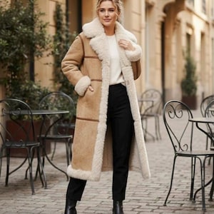 Könnte beinhalten: Ein langer, hellbrauner Shearling-Mantel mit cremefarbenem Futter und Besatz. Der Mantel hat einen breiten Kragen und wird über einem weißen Pullover und einer schwarzen Hose getragen. Schwarze Stiefeletten vervollständigen das Outfit. Europäische Straßenszene im Hintergrund.