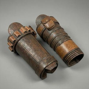 May include: Two brown leather arm guards with a distressed finish. One guard features a series of small pouches, while the other has a small flap pocket. Both are secured with straps and buckles, suggesting a historical or fantasy theme.