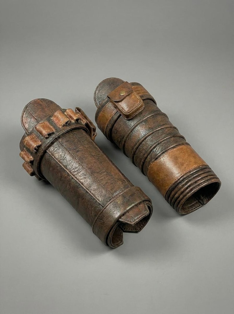 May include: Two brown leather arm guards with a distressed finish. One guard features a series of small pouches, while the other has a small flap pocket. Both are secured with straps and buckles, suggesting a historical or fantasy theme.
