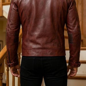 Pu&ograve; includere: Giacca in pelle bordeaux con design trapuntato su spalle e maniche. La giacca ha un colletto classico e una chiusura frontale con cerniera. La persona nell'immagine indossa pantaloni neri.