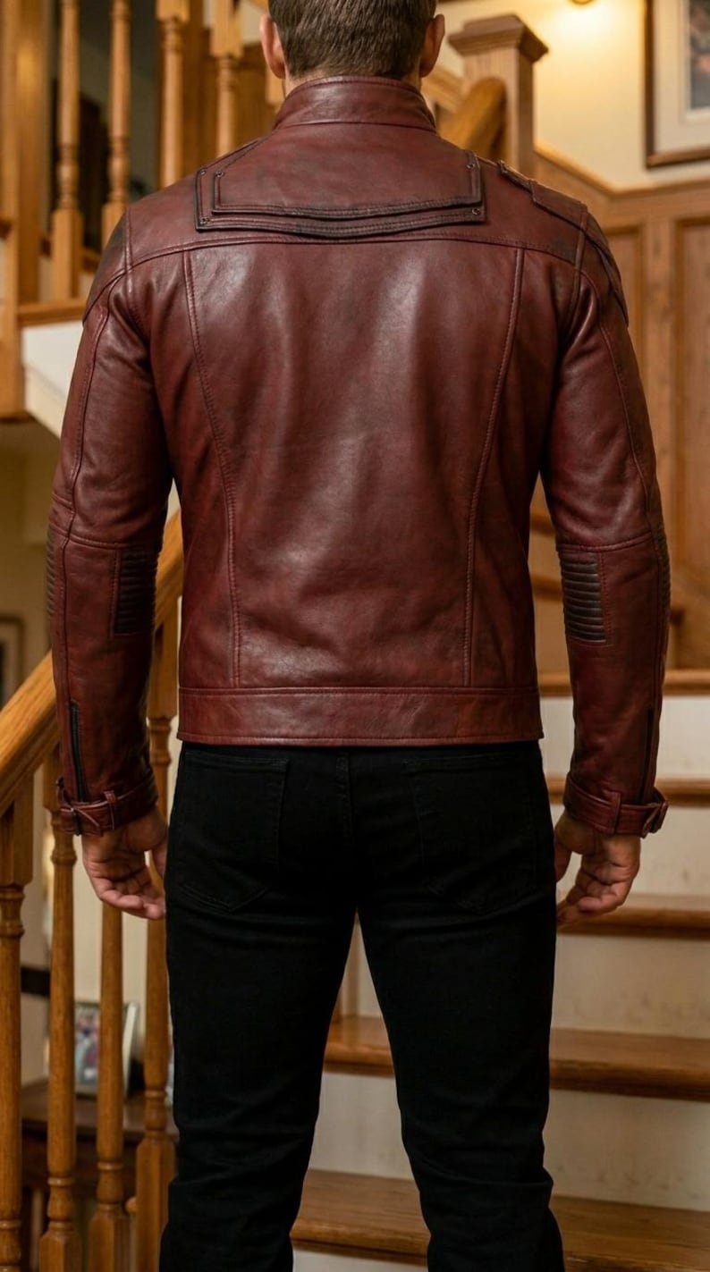 Pu&ograve; includere: Giacca in pelle bordeaux con design trapuntato su spalle e maniche. La giacca ha un colletto classico e una chiusura frontale con cerniera. La persona nell'immagine indossa pantaloni neri.