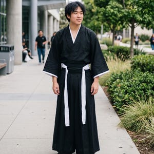 May include: A black kimono-style top with wide sleeves and white trim, paired with matching black wide-leg hakama trousers featuring a white sash tied at the waist. The outfit is completed with traditional black and beige thong-style sandals.