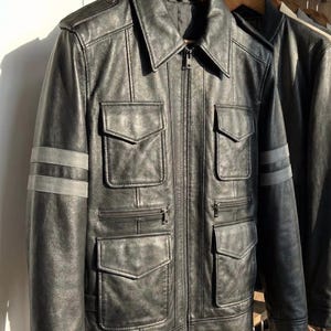 May include: Black leather jacket with a zip-up front, multiple front pockets, and gray stripes on the sleeves. The jacket is hanging on a wooden hanger. A second jacket is partially visible.