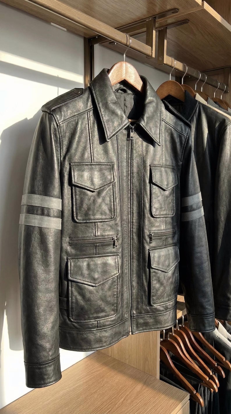 May include: Black leather jacket with a zip-up front, multiple front pockets, and gray stripes on the sleeves. The jacket is hanging on a wooden hanger. A second jacket is partially visible.