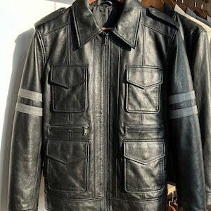 May include: Black leather jacket with a zip-up front, multiple front pockets, and gray stripes on the sleeves. The jacket is hanging on a wooden hanger. A classic outerwear piece.