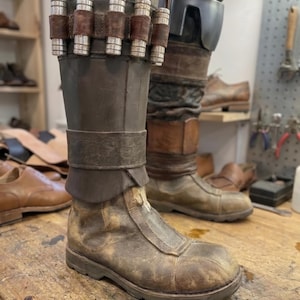 May include: A pair of weathered brown leather boots with a worn, distressed appearance. The boots feature a high shaft, a strap detail, and metal accents. The boots are on a wooden surface, suggesting a workshop setting.