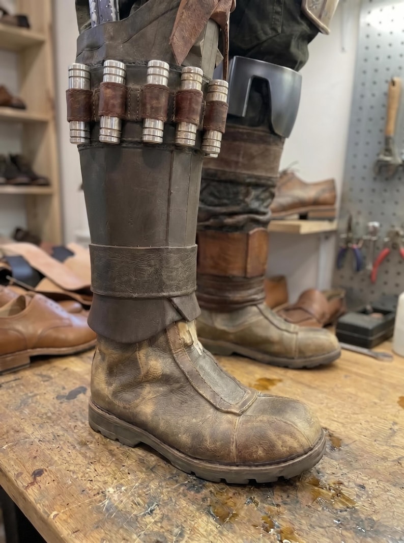 May include: A pair of weathered brown leather boots with a worn, distressed appearance. The boots feature a high shaft, a strap detail, and metal accents. The boots are on a wooden surface, suggesting a workshop setting.