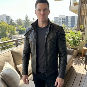 May include: A black leather jacket with a unique design, featuring textured panels on the shoulders and sleeves. The jacket has a zip-up front and a classic collar. The person is wearing a gray t-shirt and black pants.