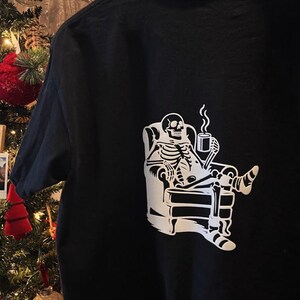 May include: Black t-shirt featuring a white graphic of a skeleton relaxing in an armchair while holding a mug of coffee. The skeleton is depicted with detailed bone structure and is seated in a comfortable pose. The shirt is made of a soft material.