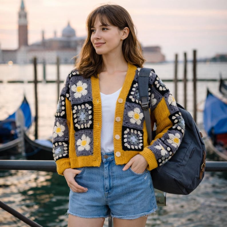 K&ouml;nnte beinhalten: Gelb, grau und wei&szlig; geh&auml;kelte Strickjacke mit einem G&auml;nsebl&uuml;mchenmuster. Die Strickjacke hat einen V-Ausschnitt, einen Knopfverschluss und lange &Auml;rmel. Das Model tr&auml;gt Jeansshorts und ein wei&szlig;es Oberteil. Ein dunkelgrauer Rucksack ist zu sehen.