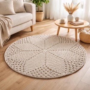 Op de afbeelding: Een ronde, crèmekleurige gehaakte vloerkleed met een geometrisch patroon. Het kleed is gemaakt van dik garen en heeft een textuur. Het ligt op een houten vloer in een woonkamer.
