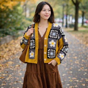 K&ouml;nnte beinhalten: Senfgelbe H&auml;keljacke mit floralem Granny-Square-Design. Die Strickjacke hat eine Knopfleiste vorne und ist mit grauen und schwarzen Quadraten akzentuiert. Das Blumenmuster enth&auml;lt wei&szlig;e und hellblaue Details. Die Person tr&auml;gt ein braunes Kleid.