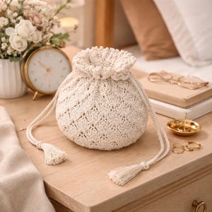 Puede incluir: Un bolso con cordón de punto de color crema con un patrón de diamantes texturizado. El bolso tiene una parte superior con volantes y dos borlas de color crema. El bolso está sobre una superficie de madera junto a un despertador dorado y un jarrón de flores.