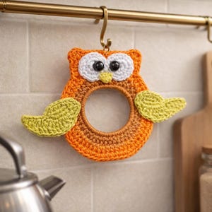 May include: A handmade crocheted owl-shaped kitchen towel holder. The owl is in shades of orange, brown, white, and yellow. It features large black eyes, white eye surrounds, and yellow wings. It hangs from a brass hook.