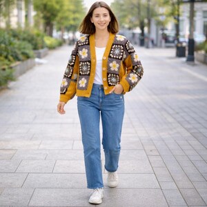 K&ouml;nnte beinhalten: Eine Frau tr&auml;gt eine senfgelbe H&auml;keljacke mit floralen Quadraten, kombiniert mit einer blauen Jeans und wei&szlig;en Turnschuhen. Die Strickjacke hat eine Knopfleiste und lange &Auml;rmel, mit einem wei&szlig;en T-Shirt darunter.