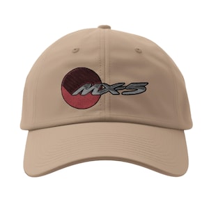 May include: A tan baseball cap featuring the embroidered text "MX-5" in a stylized font. The design includes a circular emblem with red and maroon segments. The cap has a curved brim and a fabric strap closure.