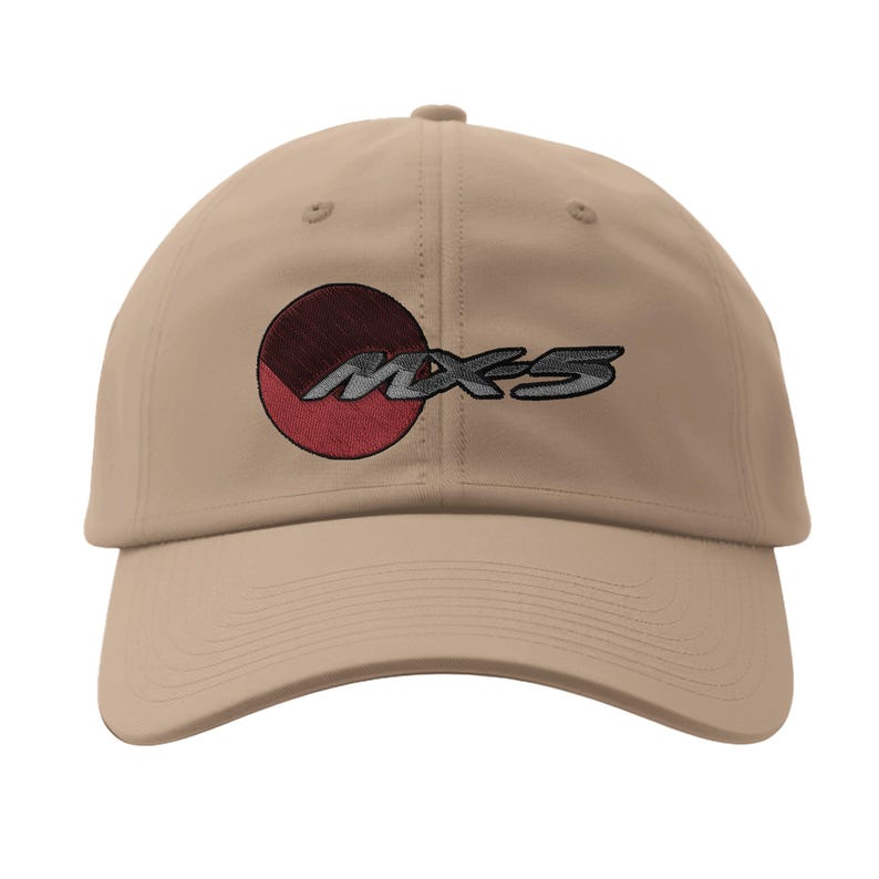 May include: A tan baseball cap featuring the embroidered text "MX-5" in a stylized font. The design includes a circular emblem with red and maroon segments. The cap has a curved brim and a fabric strap closure.