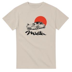 May include: A light beige t-shirt featuring a black and white illustration of a car with pop-up headlights, a red sun, and the word "Miata" in a stylized font. The design is centered on the front of the shirt.