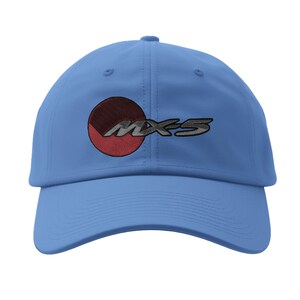 May include: A light blue baseball cap with the embroidered text "MX-5" in silver and a red and brown circle design. The cap has a curved brim and a fabric strap closure.