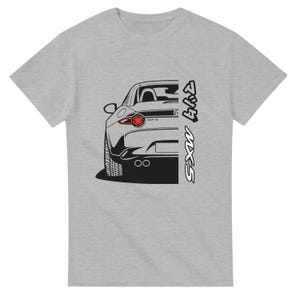 Pode incluir: T-shirt cinza claro com uma ilustração a preto e branco da traseira de um carro. O carro tem uma luz traseira vermelha e tubos de escape duplos. O texto "MX-5" é impresso verticalmente no lado direito.