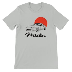 May include: A light gray short-sleeve t-shirt featuring a black line-art illustration of a sports car positioned in front of a solid red circle. Below the car, the word Miata is printed in a black, stylized, cursive-inspired script font.