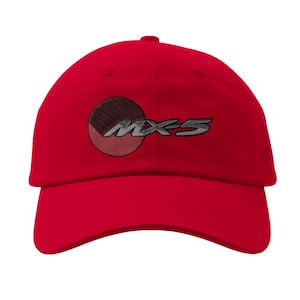 May include: A bright red baseball cap featuring the embroidered text "MX-5" in silver. The design includes a circular emblem with brown and red segments. The cap has a curved brim and a classic six-panel construction.