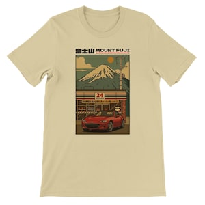 May include: Beige t-shirt with a graphic depicting Mount Fuji, a red sports car, and a convenience store with Japanese text. The design includes the text "Mount Fuji" and "24 Hours". The shirt is made of a soft material.