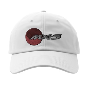 Puede incluir: Gorra de béisbol blanca con el texto bordado "MX-5" en gris, acompañado de un diseño circular rojo y granate. La gorra tiene una visera curva y una construcción clásica de seis paneles.