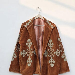 May include: A brown corduroy jacket with embroidered floral designs. The jacket has a striped interior and is hanging on a wooden hanger. The embroidery is a light beige colour.