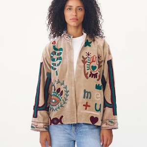 Puede incluir: Chaqueta beige bordada con símbolos y texto coloridos. La chaqueta tiene cuello alto y mangas largas con rayas decorativas. El texto "me + u" está bordado en la parte delantera. La parte inferior de la imagen tiene el texto "summerfashiongifts".