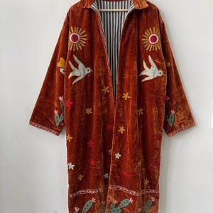 May include: A long, rust-coloured velvet coat featuring embroidered motifs of suns, birds, cacti, and small flowers. The garment has a collared neckline, long sleeves, and an interior lining with a black and white vertical stripe pattern.