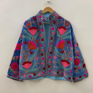 Puede incluir: Chaqueta azul con bordados florales en tonos rosa, rojo y naranja. La chaqueta tiene cuello mandarín y está forrada con una tela a rayas azul claro. El bordado presenta flores y hojas estilizadas.