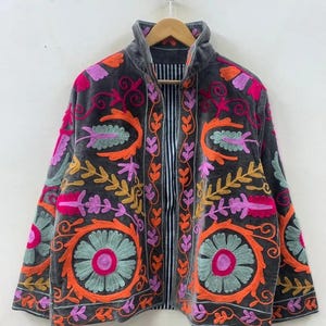 May include: A dark grey jacket with colourful floral embroidery in orange, pink, yellow, and green. The jacket has a high collar and a striped interior. The embroidery features large, circular flower designs and intricate leaf patterns.