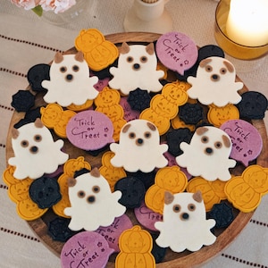 May include: A wooden platter filled with assorted Halloween-themed biscuits, including white ghost-shaped biscuits with brown ears, orange pumpkin-shaped biscuits, black skull-shaped biscuits, and round purple biscuits with the text Trick or Treat.