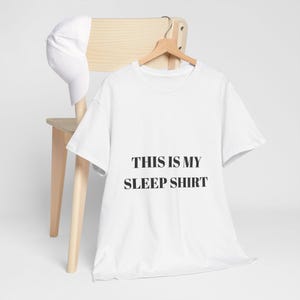 Könnte beinhalten: Ein weißes kurzärmeliges T-Shirt mit dem schwarzen Text THIS IS MY SLEEP SHIRT auf der Vorderseite, präsentiert auf einem Holzkleiderbügel neben einer schlichten weißen Baseballkappe. Das T-Shirt hat einen Rundhalsausschnitt und eine Standardpassform.