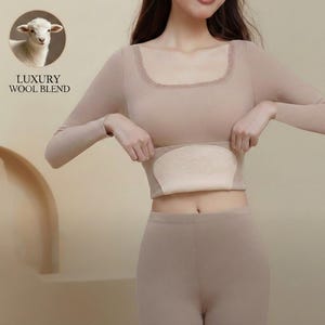 Puede incluir: Una camiseta beige de manga larga y leggings a juego. La parte superior tiene un escote cuadrado con ribete de encaje y un panel beige incorporado. La imagen incluye un logotipo que dice "LUXURY WOOL BLEND" con una ilustración de oveja.