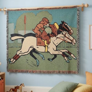 May include: A woven tapestry featuring a vintage-style illustration of two horses racing, ridden by cartoon figures. The tapestry has a green and blue background with a fringe border. The horses wear the number 10 on their saddle.