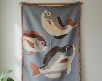 May include: A woven wall hanging featuring three stylized fish in shades of brown, beige, and orange against a light blue background. The fish are arranged on a rectangular textile with a fringed edge, suspended from a wooden rod.