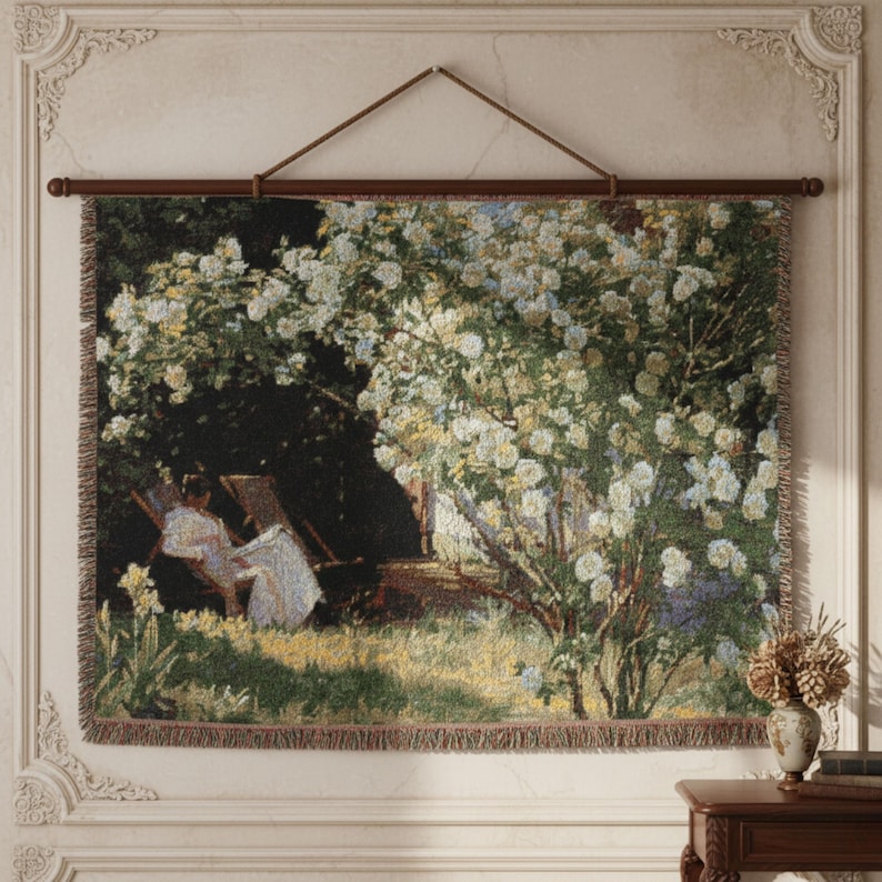 Pode incluir: Tape&ccedil;aria de parede retratando uma mulher em um vestido branco sentada em um jardim, cercada por vegeta&ccedil;&atilde;o e flores brancas. A obra de arte &eacute; emoldurada por uma barra de madeira escura e um cord&atilde;o marrom para pendurar.