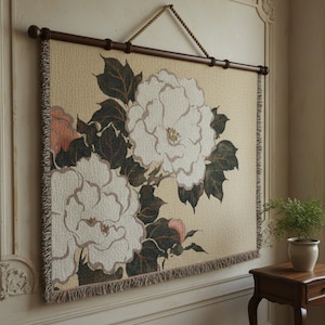 Könnte beinhalten: Ein gewebter Wandbehang mit floralem Design. Er zeigt große weiße Blüten, dunkelgrüne Blätter und Pfirsichakzente. Der Wandteppich ist an einer dunkelbraunen Holzstange befestigt und wird von einem Seil gehalten. Die Farbpalette ist neutral.
